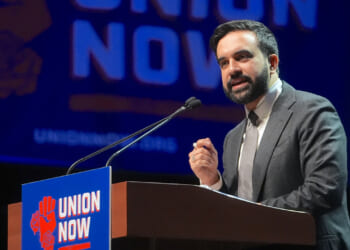 Democratic Socialists of America member and New York City Mayor Zohran Mamdani speaks behind a "union now" poster. Pictured: A man in a suit speaks at a podium with a Union Now sign, in front of a large Union Now banner.