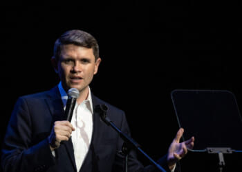 James Talarico in a suit speaks into a microphone on stage, gesturing with one hand, with a transparent teleprompter next to him against a dark background.