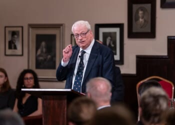 An older man in a suit speaks at a podium in a formal room with framed photos on the wall. Several people, reflecting diversity, are seated and listening attentively in the background.