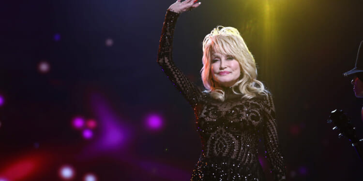 A woman in a sparkling black dress waves on stage, with colorful lights and a blurred background creating a vibrant atmosphere.
