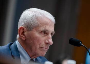 Dr. Anthony Fauci with short white hair and a blue suit sits at a desk, speaking into a microphone. The background is blurred, focusing on his serious expression.