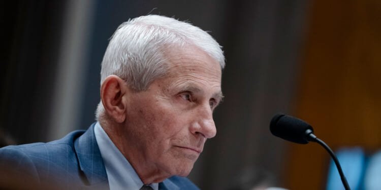 Dr. Anthony Fauci with short white hair and a blue suit sits at a desk, speaking into a microphone. The background is blurred, focusing on his serious expression.