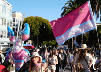 People march outdoors, some holding a large trans pride flag with a Soviet symbol. A person wears butterfly wings, and buildings, palm trees, and more flags are visible in the background. It is sunny and crowded.