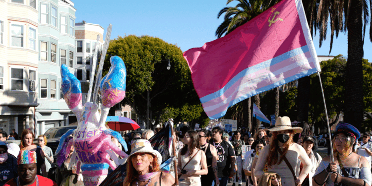 People march outdoors, some holding a large trans pride flag with a Soviet symbol. A person wears butterfly wings, and buildings, palm trees, and more flags are visible in the background. It is sunny and crowded.