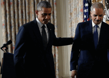 Then-President Barack Obama and then-Attorney General Eric Holder stand together indoors, one with his hand on the others back. U.S. flags and curtains are in the background. Another person claps in the background on Sept. 25, 2014.
