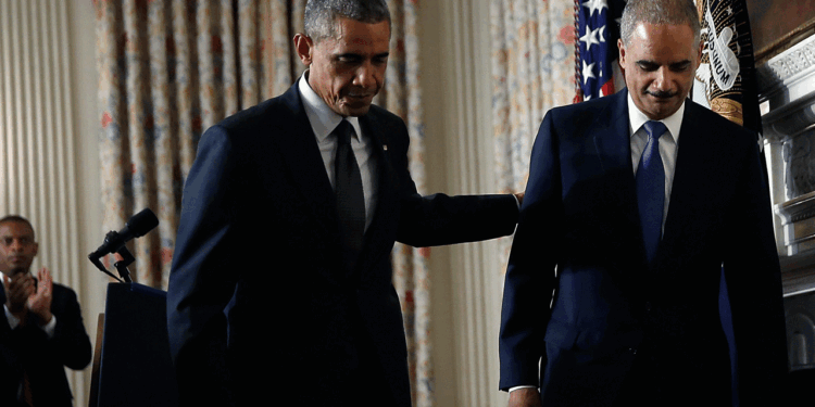 Then-President Barack Obama and then-Attorney General Eric Holder stand together indoors, one with his hand on the others back. U.S. flags and curtains are in the background. Another person claps in the background on Sept. 25, 2014.