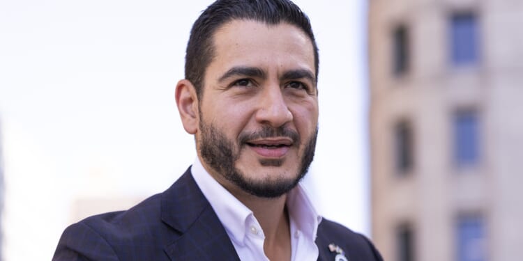 A man with short dark hair and a trimmed beard wearing a dark plaid suit jacket and white shirt stands outdoors, with blurred buildings in the background.