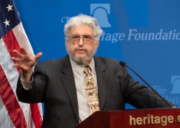 An older man with gray hair and a beard speaks at a podium with a microphone, gesturing with one hand. An American flag and a Heritage Foundation logo are visible in the background.