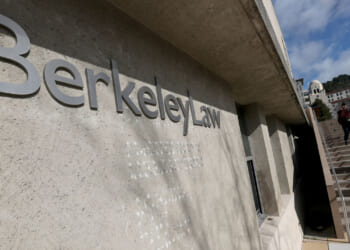 The exterior of Berkeley Law, with large metallic letters on a concrete wall and people walking up adjacent outdoor stairs on a sunny day.