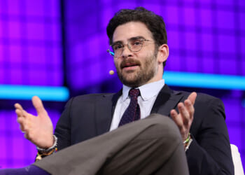 Hasan Piker responded to outrage over his singing the song "We are Charlie Kirk." Pictured: a man in a suit and glasses, wearing a headset microphone, gestures with his hands while speaking on stage against a bright purple and blue geometric background.