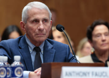 Dr. Fauci, an older man in a blue suit, sits at a table with a microphone, looking serious. There are water bottles and a nameplate in front of him. People are seated and listening in the background.