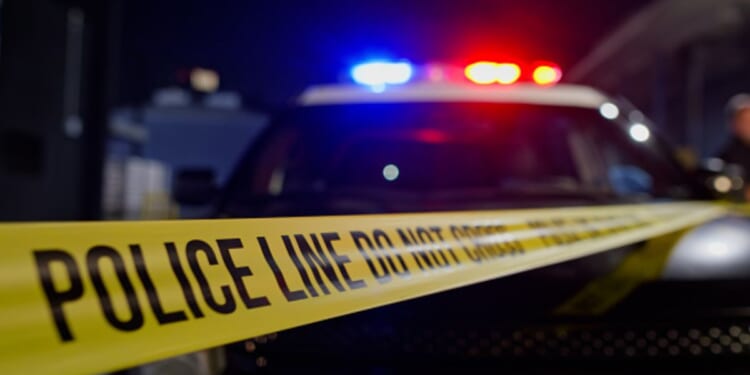 A crime scene is visible with the police lights flashing on the top of a car behind police tape.