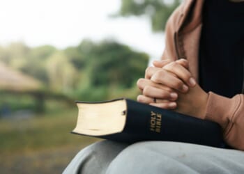 A brown woman prays with the Holy Bible in her lap outside.