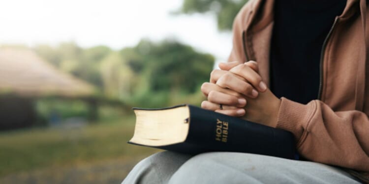 A brown woman prays with the Holy Bible in her lap outside.