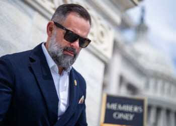 Cory Mills with a beard and sunglasses stands outside a marble building, wearing a dark suit and white shirt. A Members Only sign and a domed structure are visible in the background.