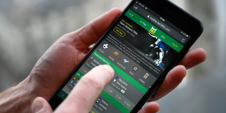 A person uses a smartphone to access a sports betting app, showing various sports icons and betting options on the screen.