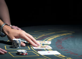 A person’s hand places chips on a blackjack table, highlighting the thrill of gambling while holding cards next to poker chips. Another person’s arm is nearby, and playing cards are visible on the felt.