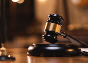 A gavel lays on a wooden block on a wooden table in a dimly lit room.