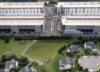 Aerial view of a data center with many parked cars, situated behind a green landscape with trees and several suburban houses at the bottom of the image.