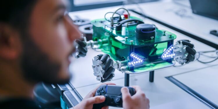A Siemens trainee holds a game controller, operating a green robotic vehicle with LED lights and mecanum wheels on a table in a modern lab.