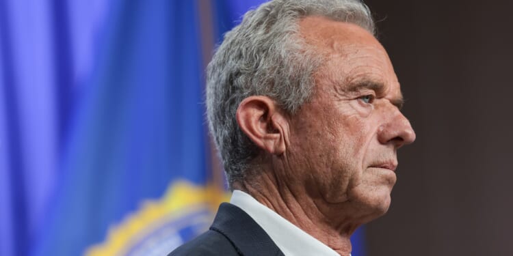 A man with gray hair in a suit stands in profile against a blue background with a blurred emblem or flag.