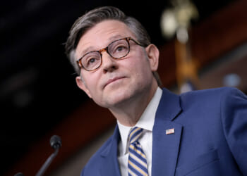 House Speaker Mike Johnson. Pictured: a man in a blue suit, striped tie, and glasses stands at a podium. An American flag pin is on his lapel, and a blurred flag is in the background.