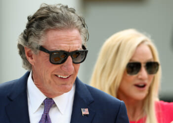 Doug Bergum in a blue suit and sunglasses smiles, with a small U.S. flag pin on his lapel. A blonde woman in sunglasses stands behind him, partially out of focus, wearing a red top.