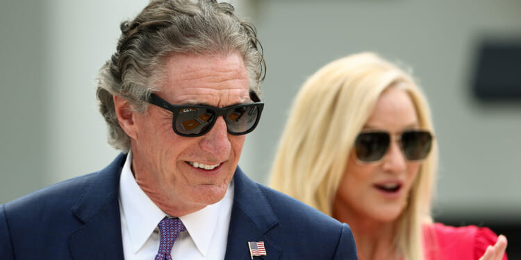 Doug Bergum in a blue suit and sunglasses smiles, with a small U.S. flag pin on his lapel. A blonde woman in sunglasses stands behind him, partially out of focus, wearing a red top.
