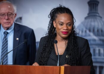 A woman smiles at a podium with microphones, wearing a black suit. An older man in a suit stands behind her. A blurred government building is in the background.