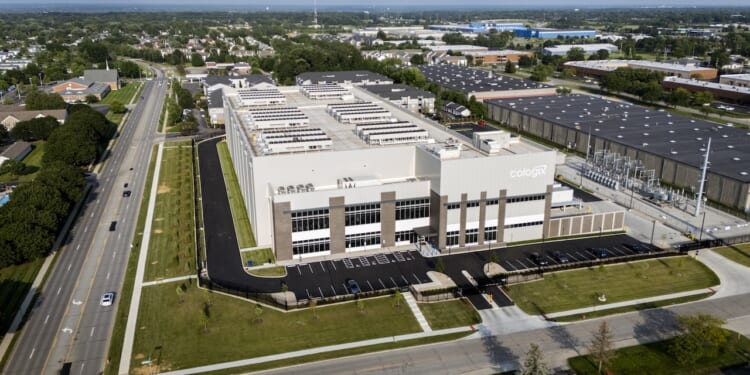 Aerial view of a large, modern data center building with the Cologix logo, surrounded by parking lots, roads, trees, and nearby industrial and residential areas.