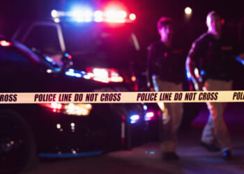 Police tape in the foreground in front of two officers and a police car with flashing lights at night.