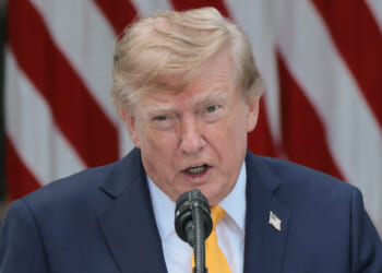 Donald Trump in a navy suit and yellow tie speaks into a microphone, with red and white striped flags in the background.