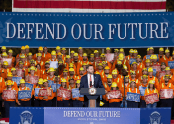 Vice President JD Vance stands at the podium as he addresses a crowd of construction workers in orange gear, with signs reading Defend Our Future and Invest in America. A large banner behind reads DEFEND OUR FUTURE.