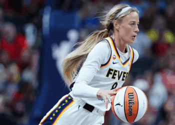 A basketball player in a white Fever uniform dribbles a basketball during a game, with focus and intensity, as the crowd watches in the blurred background.