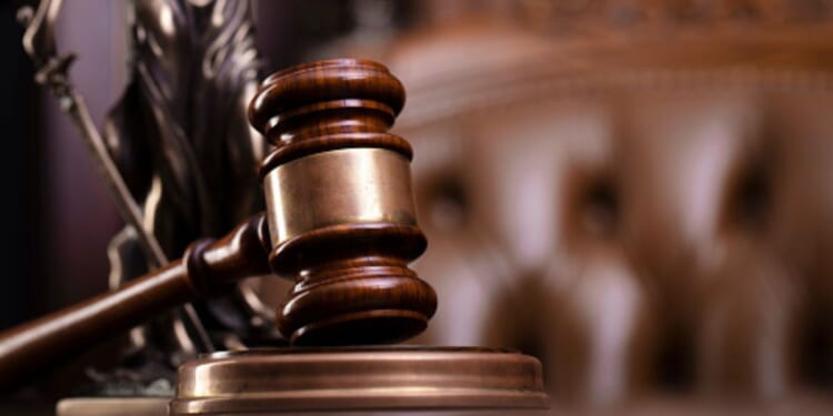 A gavel lined with gold sits on a wooden block ahead of a leather chair.