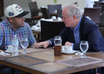 Kilmar Abrego Garcia and Sen. Chris Van Hollen sit at a restaurant table having a serious conversation. Garcia in a plaid shirt and cap, faces Van Hollen, who is dressed in a suit. Water glasses, cups, and a water bottle are on the table. The background shows empty chairs and tables.