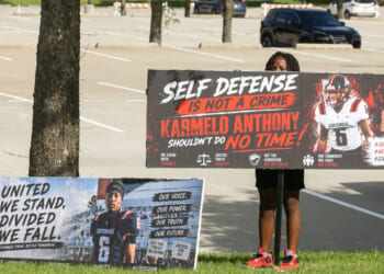 A person holds a large sign reading, “Self defense is not a crime. Karmelo Anthony shouldn’t do no time!” Another sign on the ground reads, “United we stand, divided we fall.” The scene unfolds in a parking lot with cars visible in the background, emphasizing public support for Karmelo Anthony.