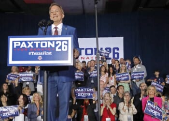 A man speaks onstage at a podium with Paxton 2026 signs, surrounded by a cheering crowd holding similar campaign signs, at what appears to be a political rally.