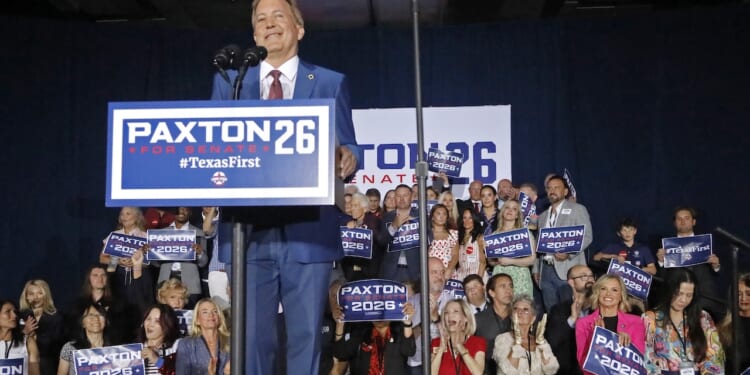 A man speaks onstage at a podium with Paxton 2026 signs, surrounded by a cheering crowd holding similar campaign signs, at what appears to be a political rally.