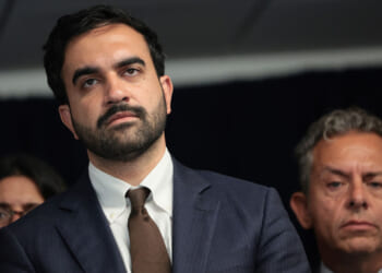 A man in a suit and tie stands in front of a dark background, looking serious. Two other people, also appearing serious, are partially visible beside and behind him.