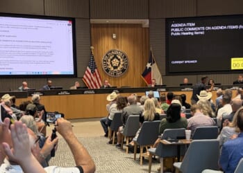 A large group attends a city council meeting. People sit facing a panel of officials at the front. Digital screens display agenda items and a timer. U.S. and Texas flags are visible behind the officials.