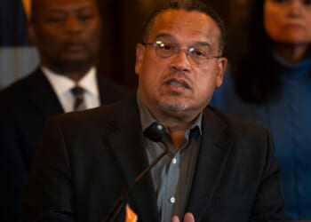 Keith Ellison glasses and a dark blazer speaks at a podium, with two people standing behind him, during an event in Minnesota.