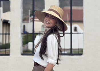 Allison Roberts is suing Portland Public schools over retaliation for her opposition to transgender lessons in school. Pictured: Roberts, a woman in a wide-brimmed straw hat and white blouse smiles while standing outdoors in front of a light-colored wall with iron-barred windows.