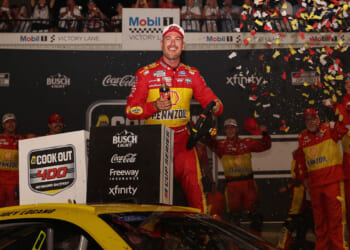 NASCAR race car driver Joey Logano in a yellow Pennzoil suit celebrates atop his car in victory lane as confetti falls. Crew members behind him cheer and clap, while racing sponsor logos cover the background.