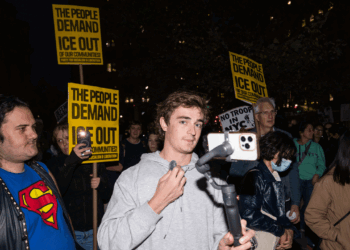 Nick Shirley speaks into a microphone connected to a phone at a nighttime protest. People around him hold yellow signs reading THE PEOPLE DEMAND ICE OUT OF OUR COMMUNITIES. The crowd includes diverse ages and attire.