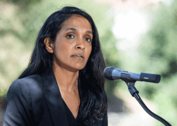 A woman with long dark hair, wearing a black blazer, stands outdoors speaking into a microphone. Greenery appears blurred in the background.