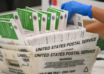A gloved hand handles a stack of mail-in ballots in green and white envelopes inside a United States Postal Service tray.