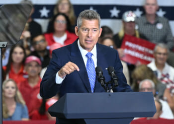 Man in a blue suit speaking at a podium with microphones, addressing a crowd in front of a patriotic backdrop featuring stars and stripes. Audience members are seated and appear to be listening.