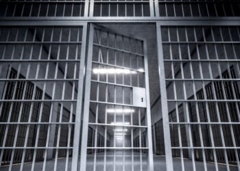 The corridor of a prison with bars and an open cell door.