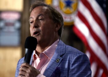 A man in a blue checkered blazer and pink shirt speaks into a microphone. An American flag and blurred background with signage are visible behind him.
